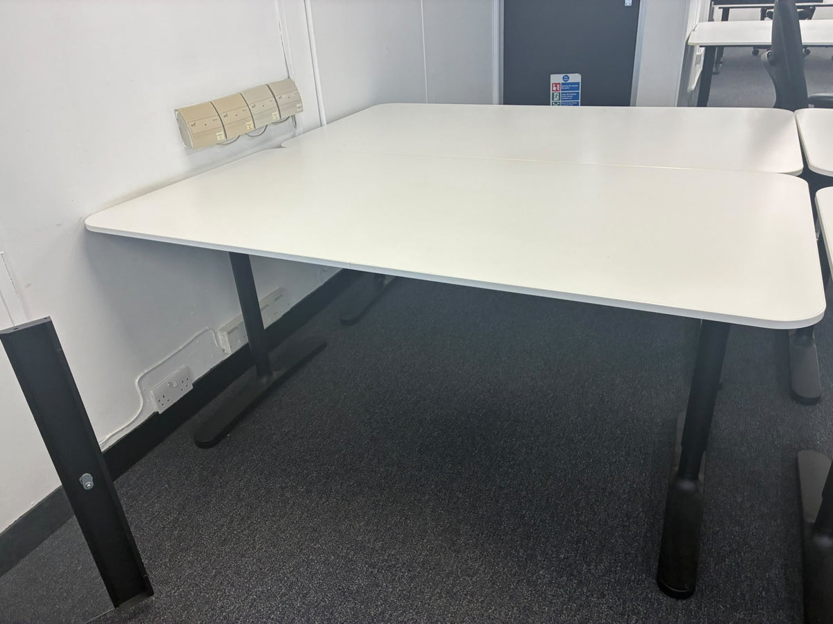 A white rectangular meeting table with black metal legs, shown in an office or classroom setting with gray carpet and a white wall.