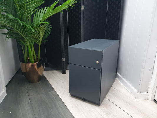 A gray slimline narrow office filing cabinet with two drawers positioned under a desk, next to a potted plant.