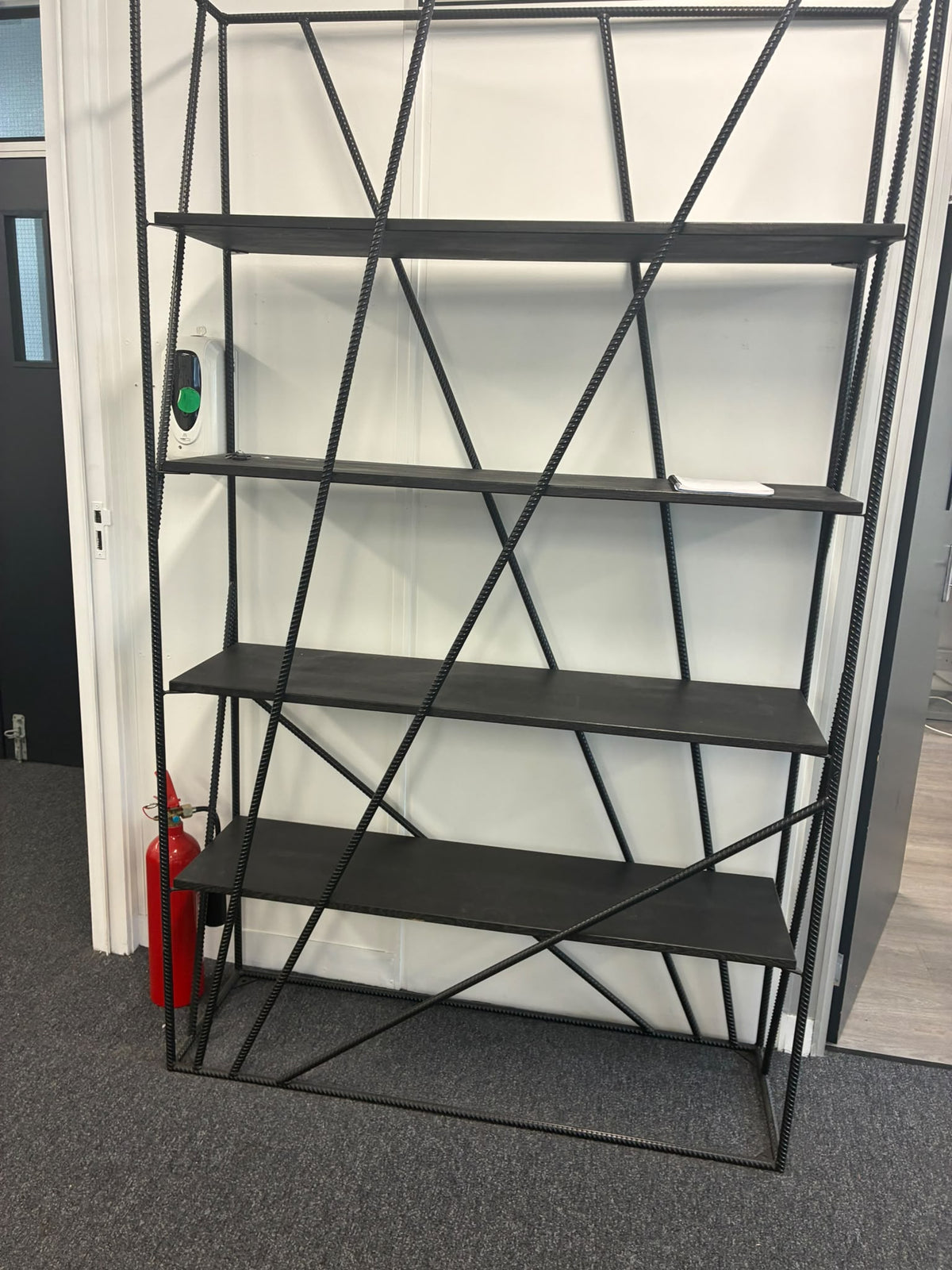 A tall black metal shelving unit with five shelves and diagonal crisscross support beams, standing in an indoor room with a white wall, gray carpet, and a red fire extinguisher visible on the left.