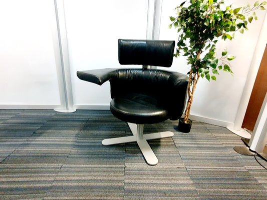 A black Kinnarps Drabert Hotspot swivel armchair with a white base, positioned in an office setting with a potted plant in the background.