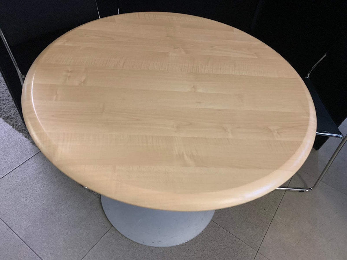 A round light wood dining table with a natural grain finish and a white pedestal base, photographed in an indoor setting.