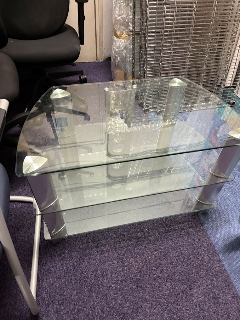 A modern two-tier glass coffee table with a clear glass top and lower shelf, supported by silver metal legs, shown in an office setting on a purple carpet.