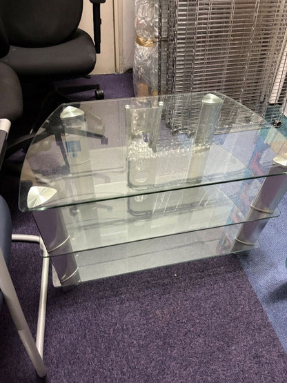 A modern two-tier glass coffee table with a clear glass top and lower shelf, supported by silver metal legs, shown in an office setting on a purple carpet.
