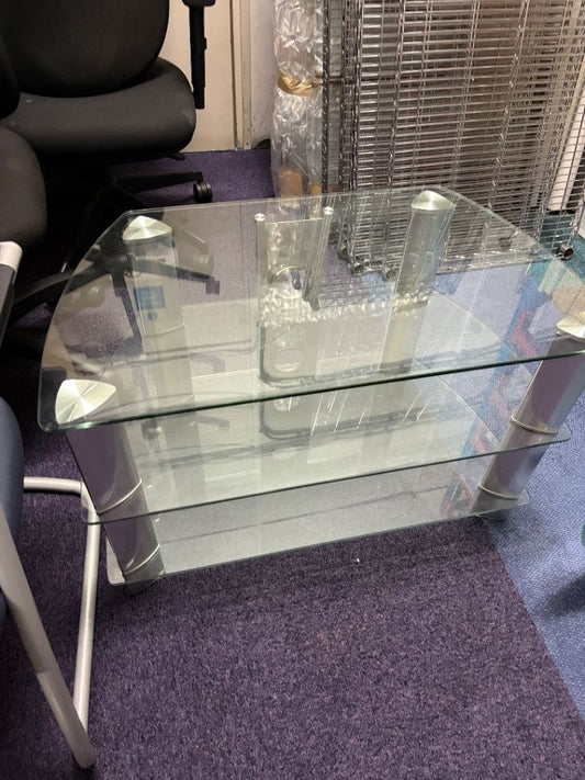 A modern two-tier glass coffee table with a clear glass top and lower shelf, supported by silver metal legs, shown in an office setting on a purple carpet.