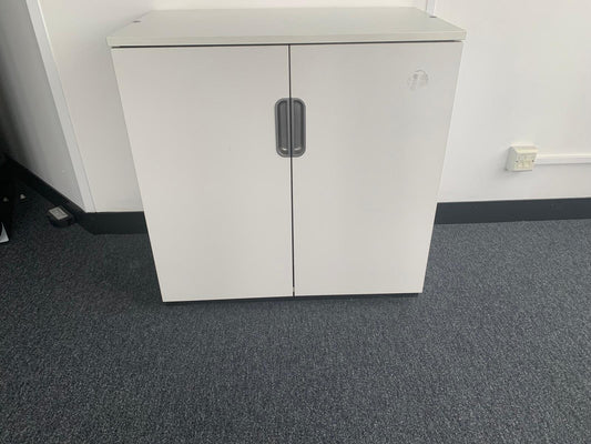 A white two-door storage cabinet with a silver handle, set against a white wall with a gray carpeted floor.