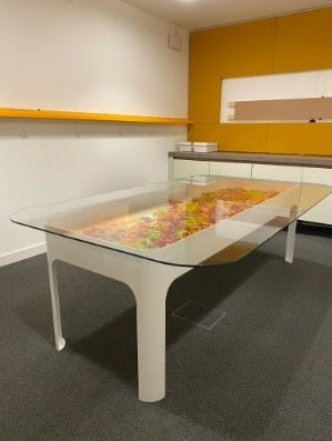 Modern white boardroom meeting conference table with glass top and colorful plastic decor inside the table, set in a room with yellow walls and a glass partition.