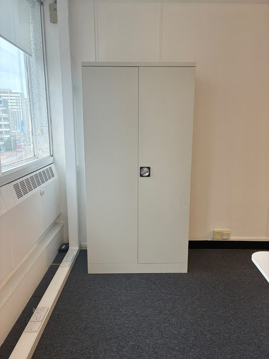 A tall, light gray two-door metal cabinet with a small lock in the center, standing in an office-like room with a gray carpet, off-white walls, and a window on the left.