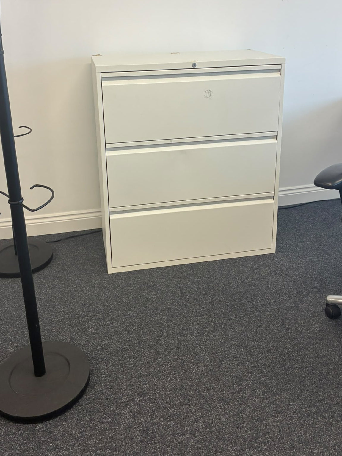 A white three-drawer metal file cabinet in an office setting, positioned against a white wall on gray carpet, with a black coat rack on the left and a black office chair on the right.
