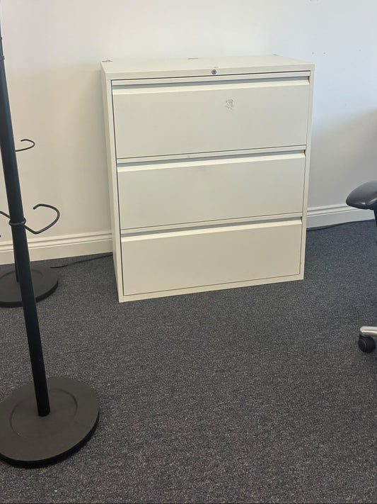 A white three-drawer metal file cabinet in an office setting, positioned against a white wall on gray carpet, with a black coat rack on the left and a black office chair on the right.
