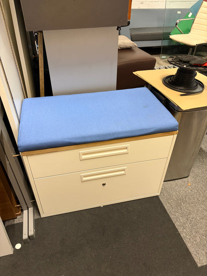 A small storage cabinet with two cream drawers and a light blue upholstered top cushion, photographed in an office-like interior with other furniture visible around it.