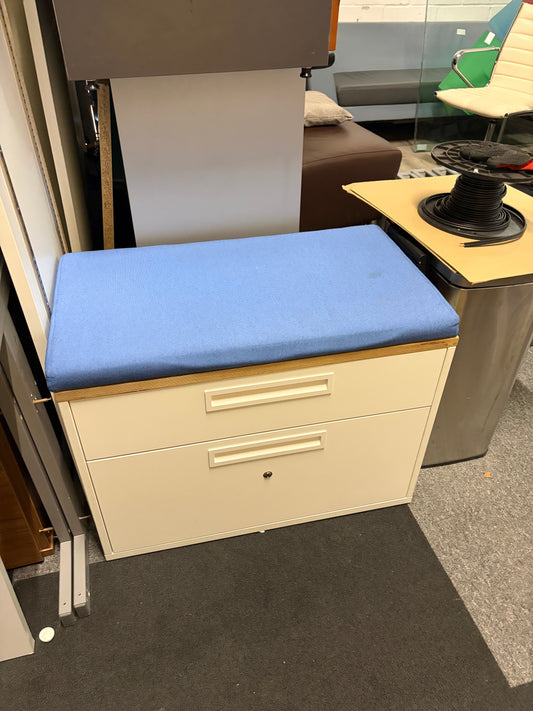 A small storage cabinet with two cream drawers and a light blue upholstered top cushion, photographed in an office-like interior with other furniture visible around it.