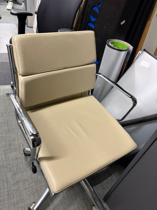 A modern office chair with a beige faux-leather seat and backrest, supported by a chrome metal frame with casters, photographed in a warehouse or showroom setting.