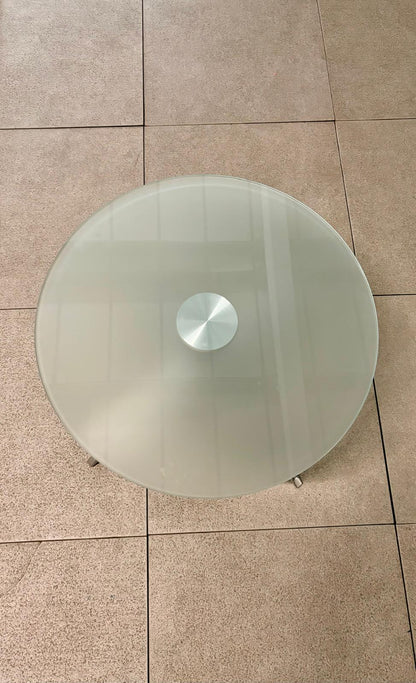 A round, transparent frosted glass tabletop with a brushed silver metal center cap, shown from a top-down perspective on a beige tiled floor.