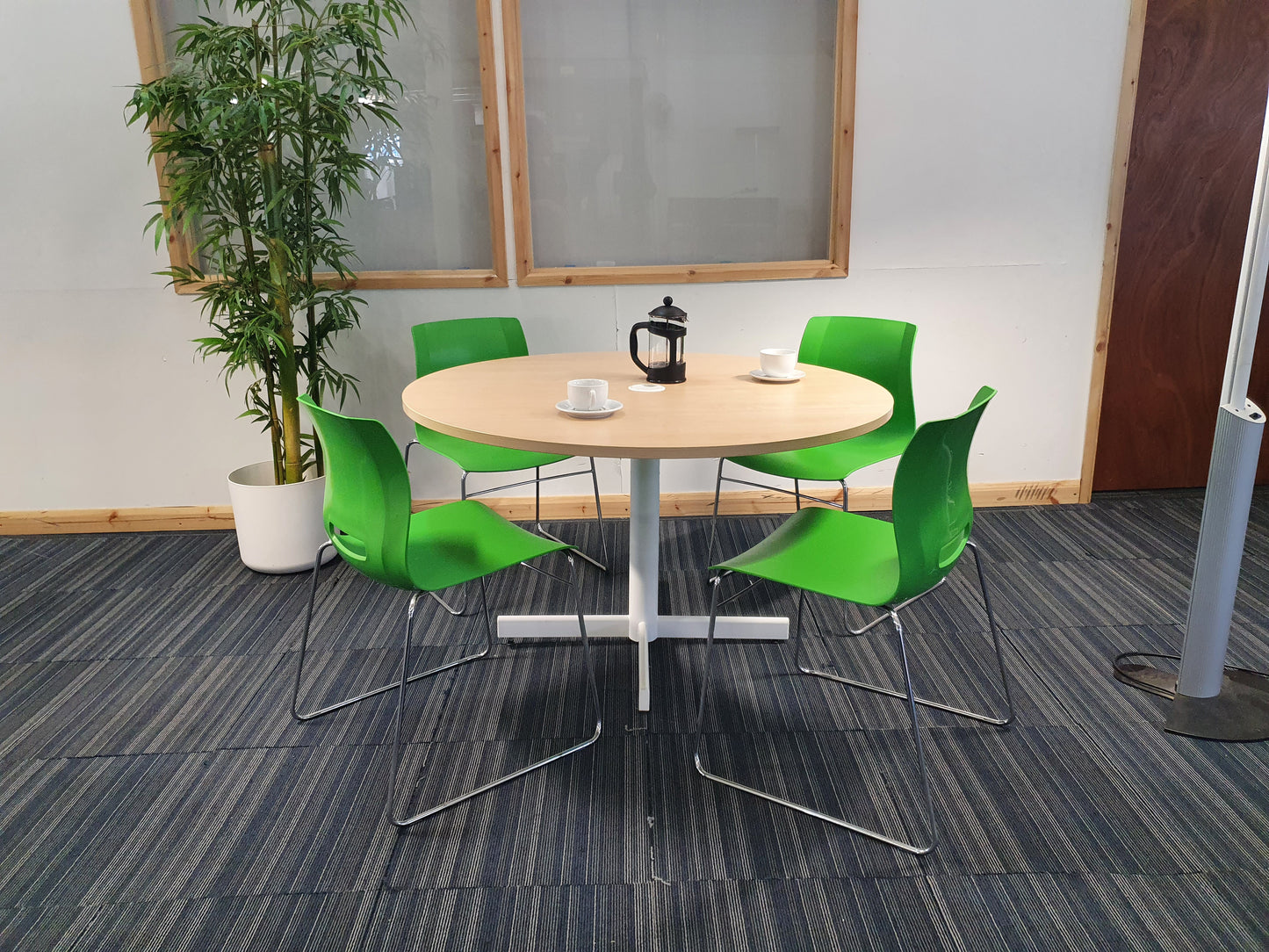 Left, tall green plant, centre, four green canteen chairs around brown round table