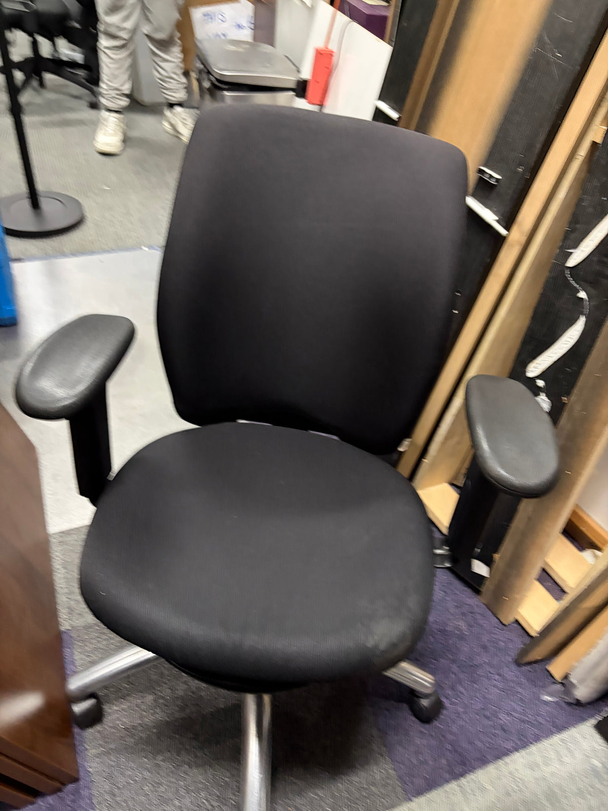A black fabric office chair with adjustable armrests and a wheeled base, photographed in a real indoor setting with other furniture and a person partially visible in the background.