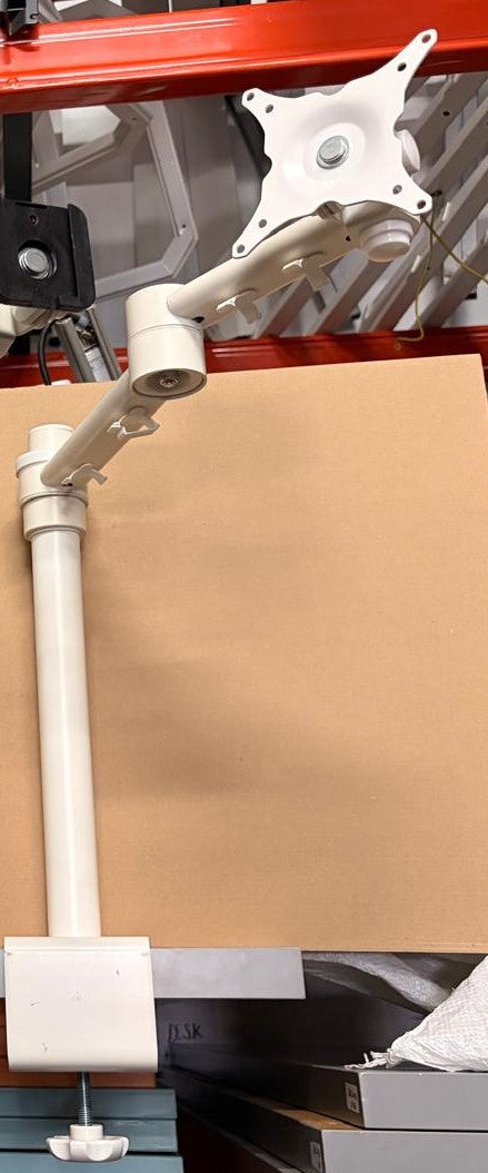 A white adjustable monitor arm with a multi-arm design and a mounting bracket, shown mounted against a large tan board in a warehouse-like setting.