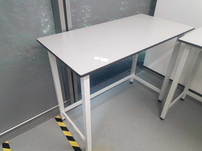 A rectangular white workbench with a white metal frame and casters, photographed in an office or workshop setting next to a glass window.