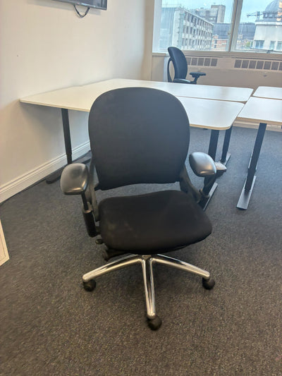 A black fabric office chair with a five-star chrome base and casters, positioned in front of a white desk in an office setting.
